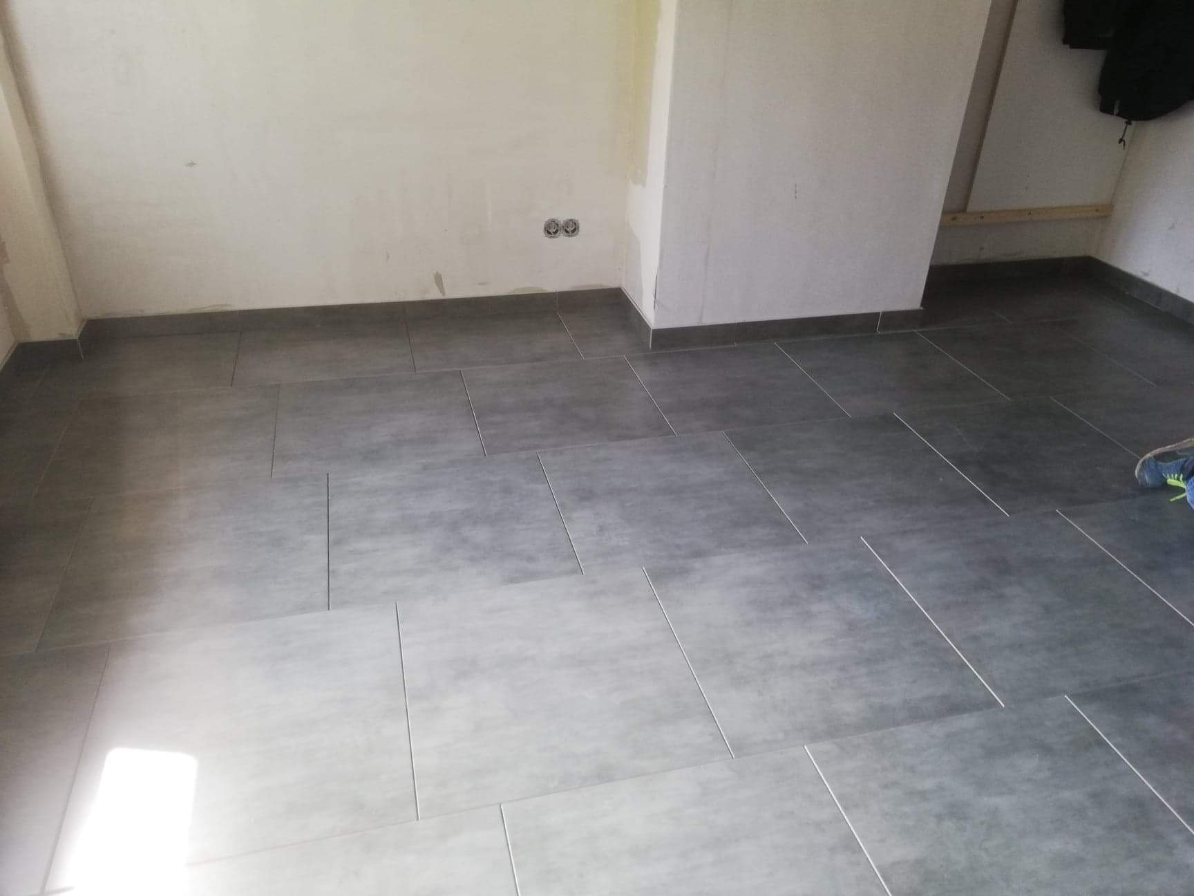 Ułożone szare płytki podłogowe w pomieszczeniu z surowymi ścianami, widoczne gniazdka elektryczne i fragment buta w prawym dolnym rogu.
