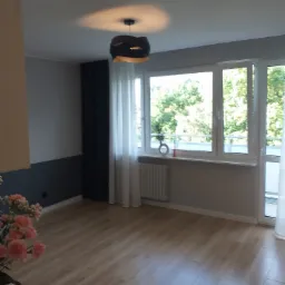 Jasne drewniane panele podłogowe w przestronnym pokoju z szarymi ścianami, ciemną zasłoną i dużym oknem z widokiem na zieleń, nowoczesna lampa sufitowa.