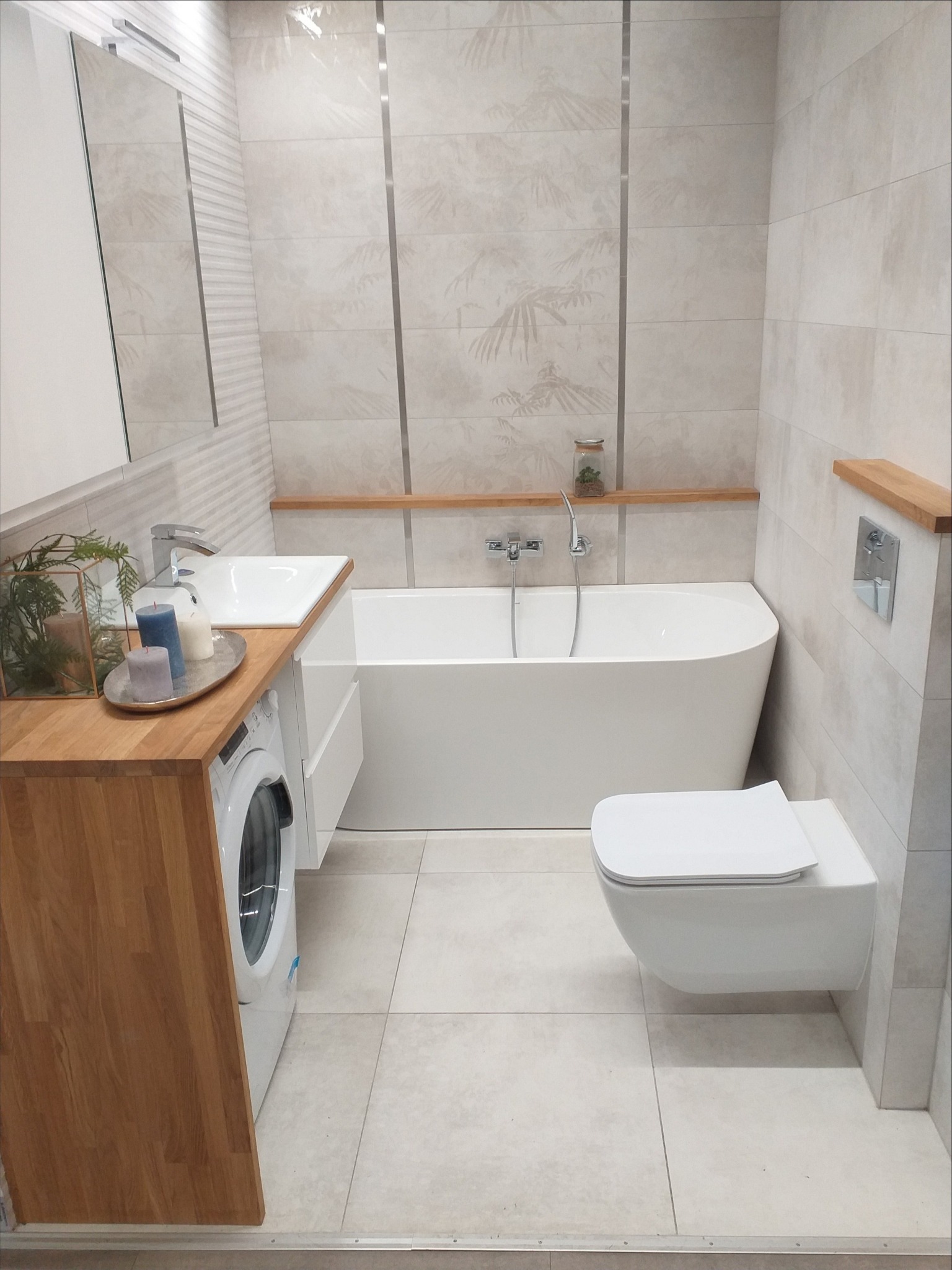 Nowoczesna łazienka z wanną, umywalką wbudowaną w drewniany blat z pralką, wiszącą toaletą i płytkami w odcieniach beżu z motywem roślinnym, uzupełniona drewnianymi półkami.