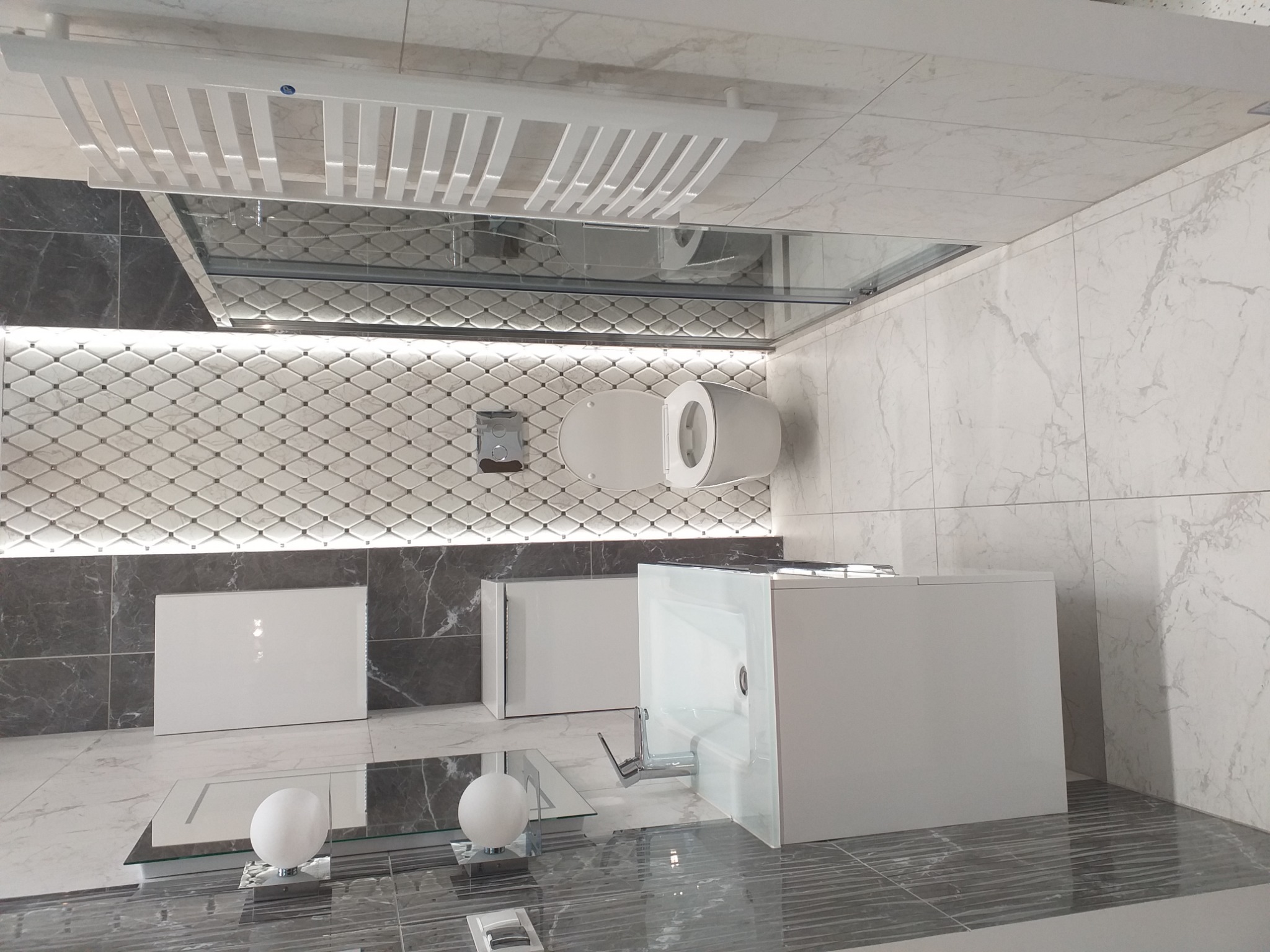 Nowoczesna łazienka z białymi płytkami imitującymi marmur, podwieszana toaleta, grzejnik drabinkowy, umywalka w zabudowie i dekoracyjna ściana z mozaiki w romby podświetlona listwą LED.