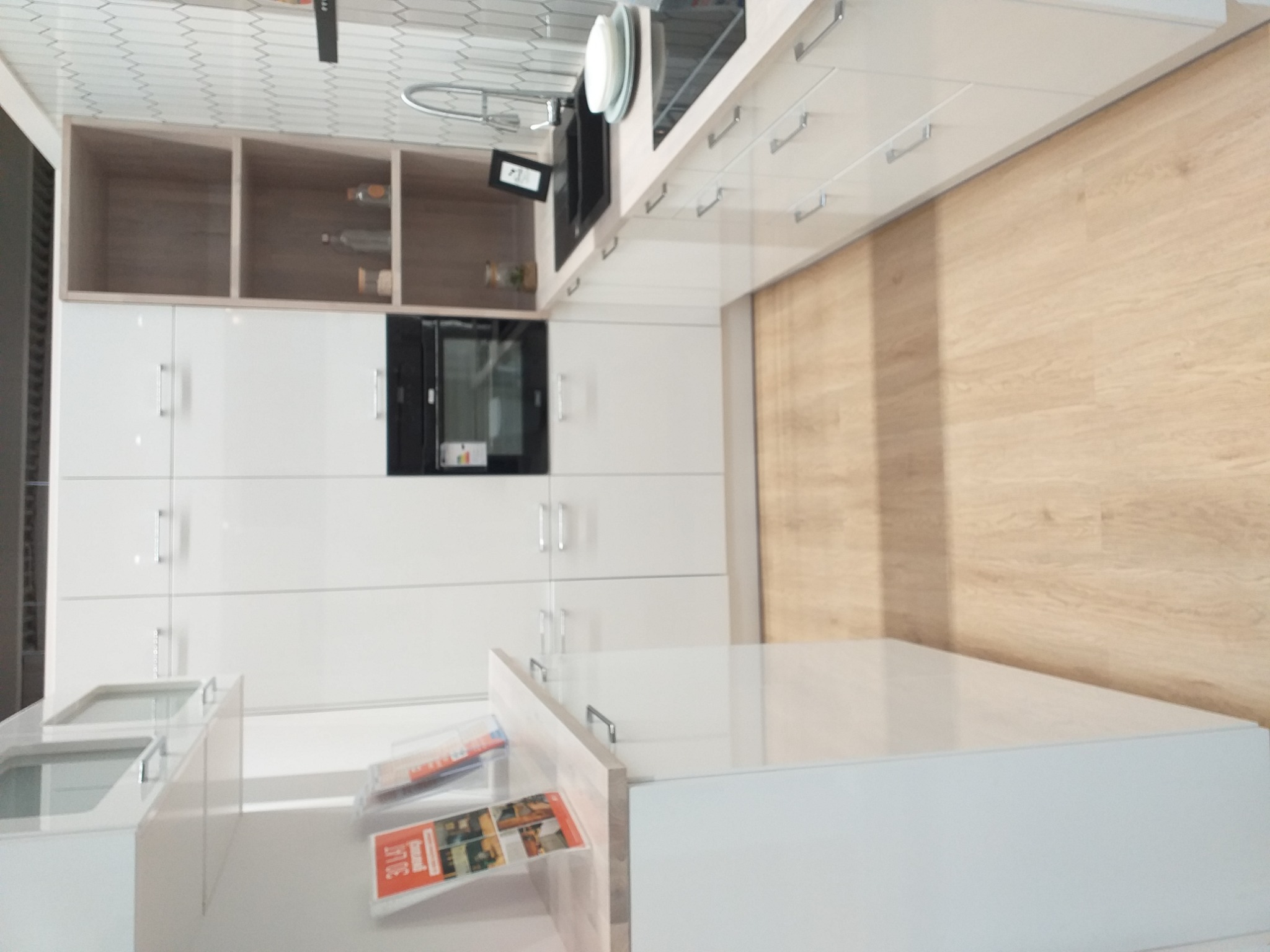 Nowoczesna, biała kuchnia z błyszczącymi frontami, drewnopodobną podłogą i zabudową z piekarnikiem oraz półkami, widoczny fragment blatu z płytą grzewczą i zlewem.