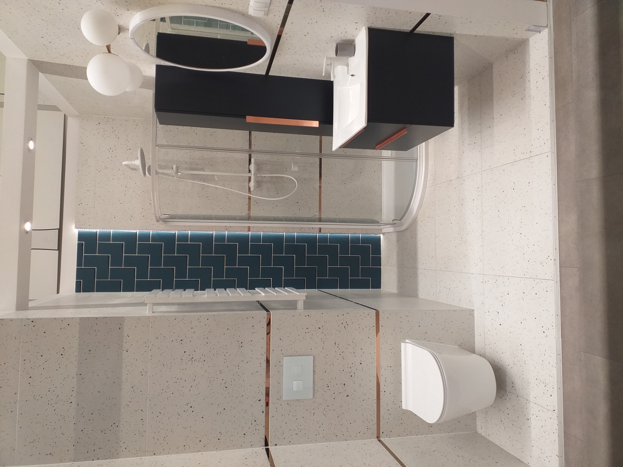 Nowoczesna łazienka z kabiną prysznicową, wiszącą szafką z umywalką, podwieszaną toaletą, lustrem z oświetleniem i płytkami imitującymi lastryko z miedzianymi akcentami.