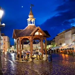 Nocny widok na Rynek Główny z drewnianą studnią pośrodku, oświetlony lampionami, z grupą ludzi siedzących wewnątrz, mokry bruk odbija światła, w tle budynki i restauracje z parasolami.