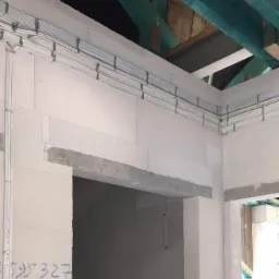 Wnętrze w trakcie budowy z widocznymi, równolegle ułożonymi przewodami elektrycznymi zamocowanymi do sufitu i ścian, surowy stan wykończenia, ściany z betonu komórkowego z niebieskim napisem.