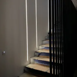 Schody w trakcie remontu z zabezpieczonymi stopniami, oświetlone trzema pionowymi liniami LED na ścianie i częściowo zasłonięte czarną, pionową balustradą.