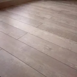 Ułożona pod kątem podłoga z paneli imitujących drewno, widoczne listwy przypodłogowe i fragment ściany z grzejnikiem w tle.