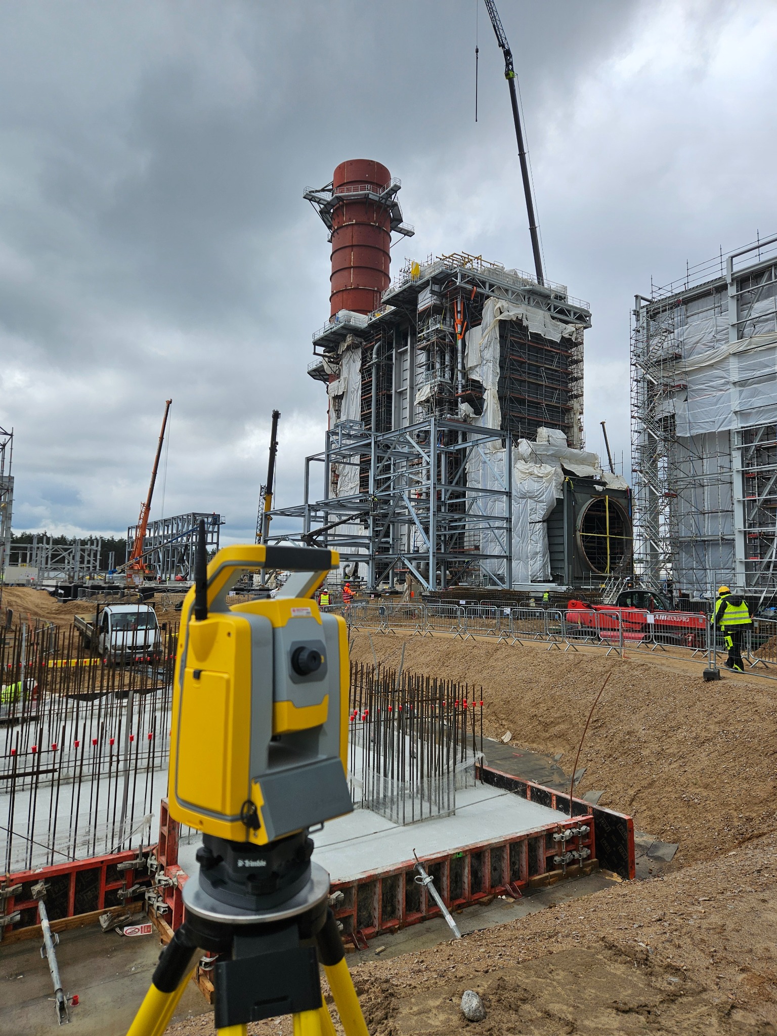 Żółty teodolit na statywie na pierwszym planie, w tle budowa dużej instalacji przemysłowej ze stalową konstrukcją, rusztowaniami i żurawiami, pochmurne niebo.