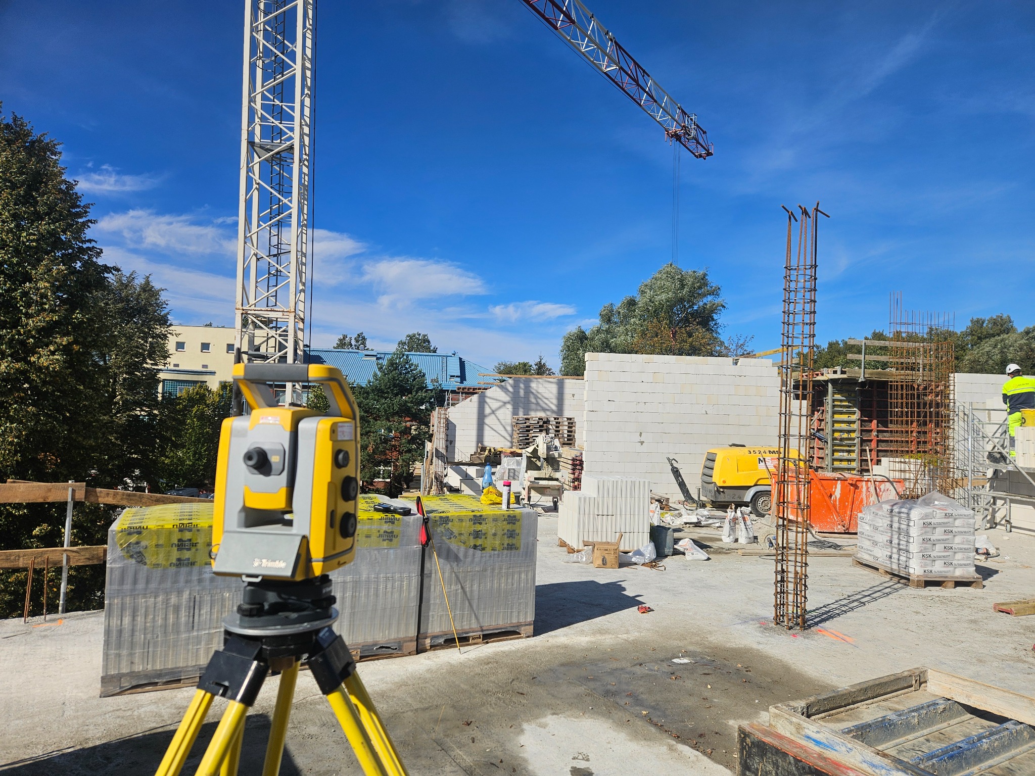 Żółty teodolit Trimble na budowie z blokami, zbrojeniami i żurawiem w tle. Prace geodezyjne w trakcie wznoszenia budynku z betonu komórkowego.