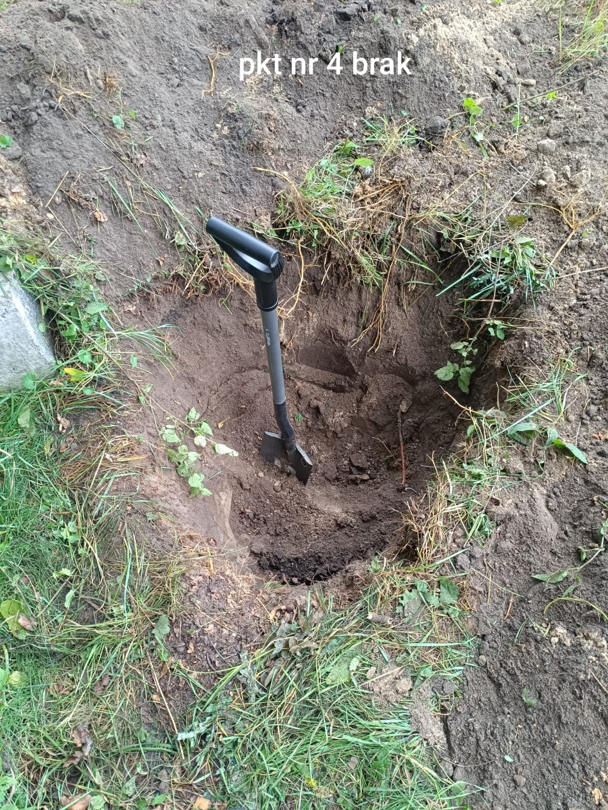 Wykonany wykop w ziemi z wbita łopatą, otoczony trawą i ziemią. Widok z góry na zagłębienie w gruncie, prawdopodobnie pod fundament lub instalację.