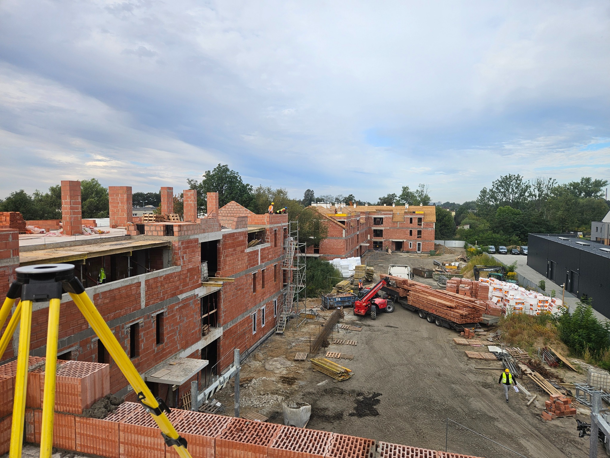 Budowa bloków z czerwonej cegły, widok z góry. Na pierwszym planie żółty statyw, w tle budynki w stanie surowym, materiały budowlane i pracownicy.