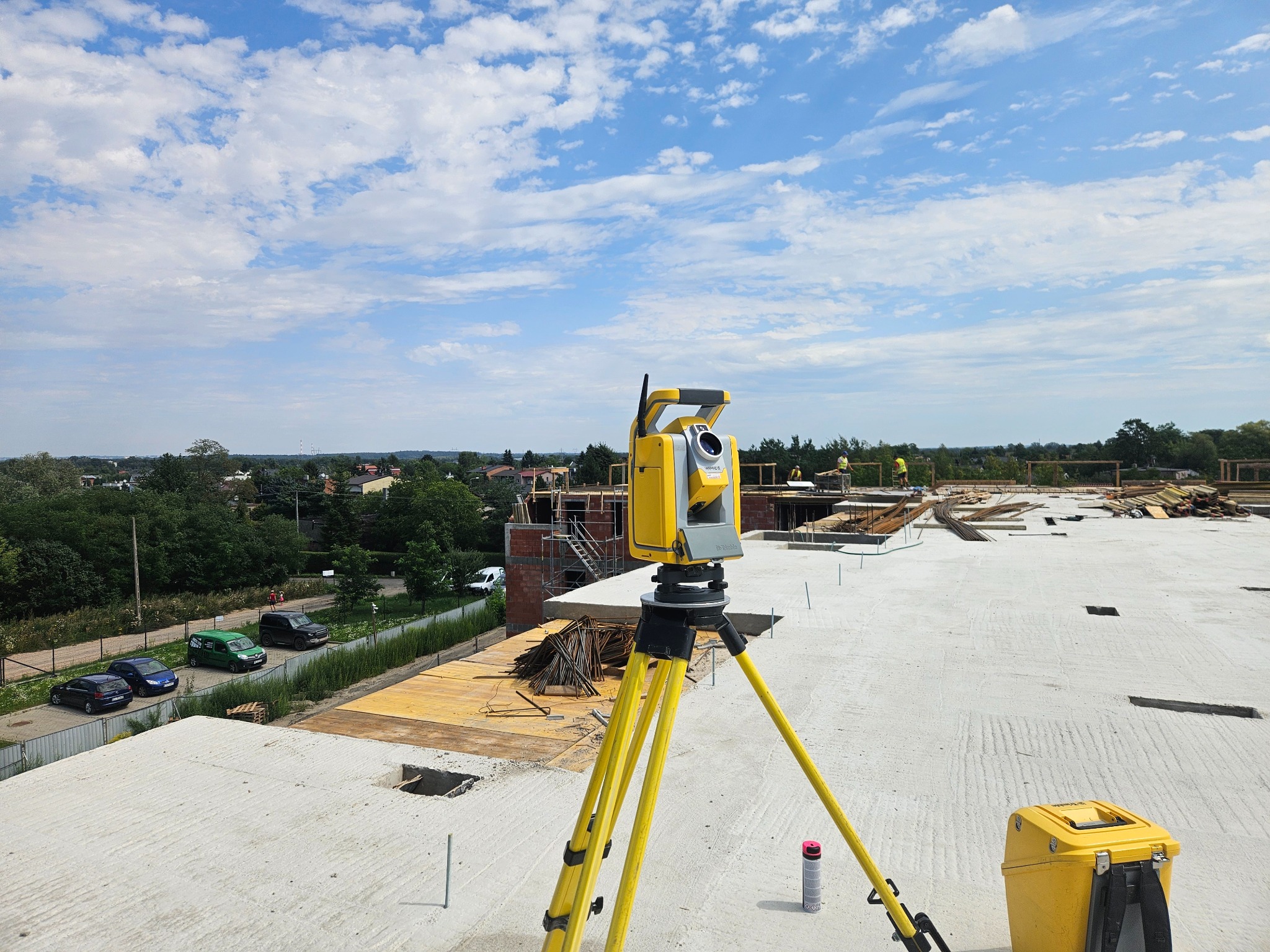 Żółty tachimetr na statywie na betonowym stropie budynku w budowie, w tle błękitne niebo z chmurami i zabudowa. Widoczne elementy konstrukcyjne i narzędzia.