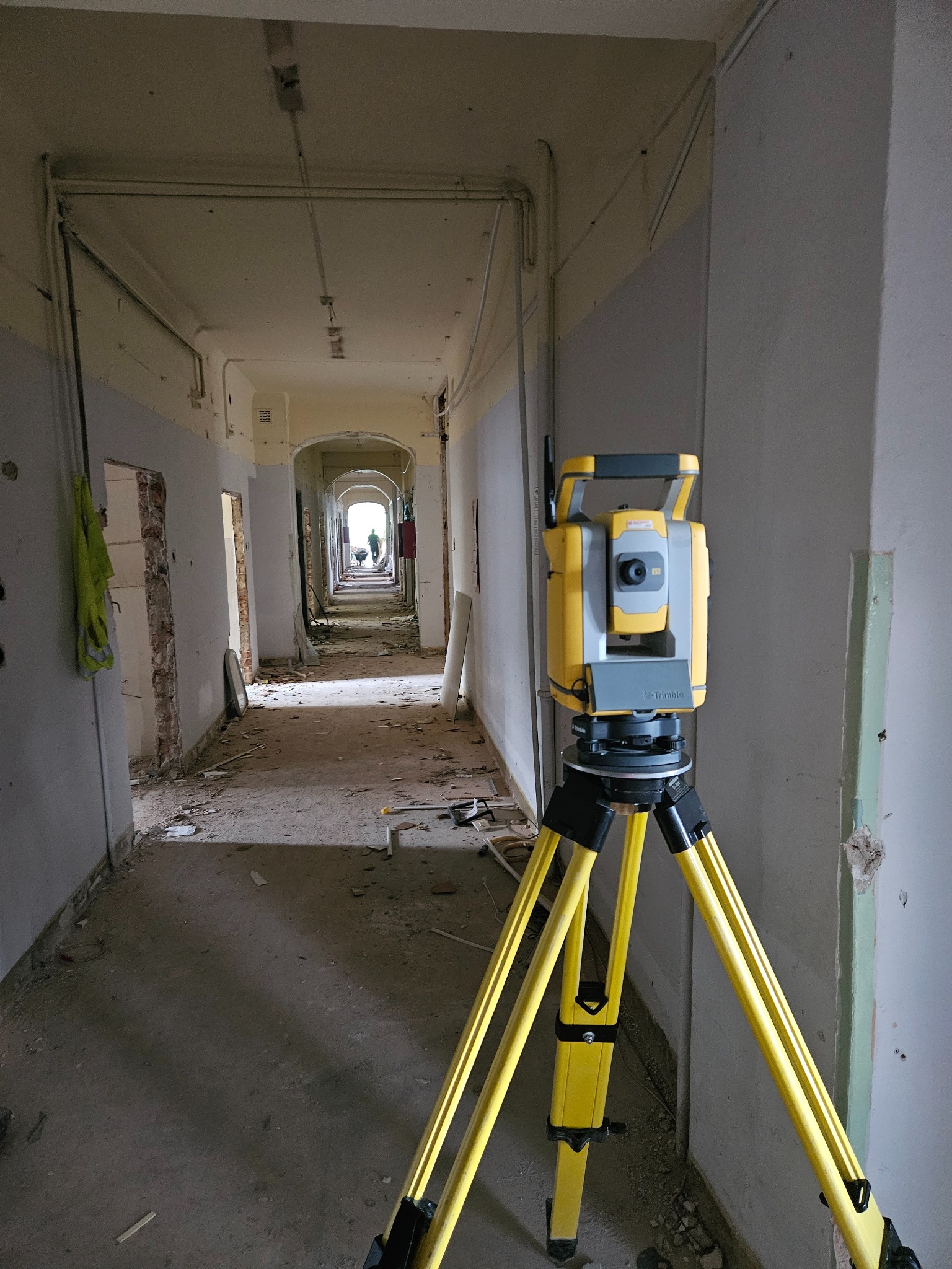 Żółty teodolit na trójnogu w korytarzu budynku w trakcie remontu. W tle osoba z taczką. Pomiar w budynku.