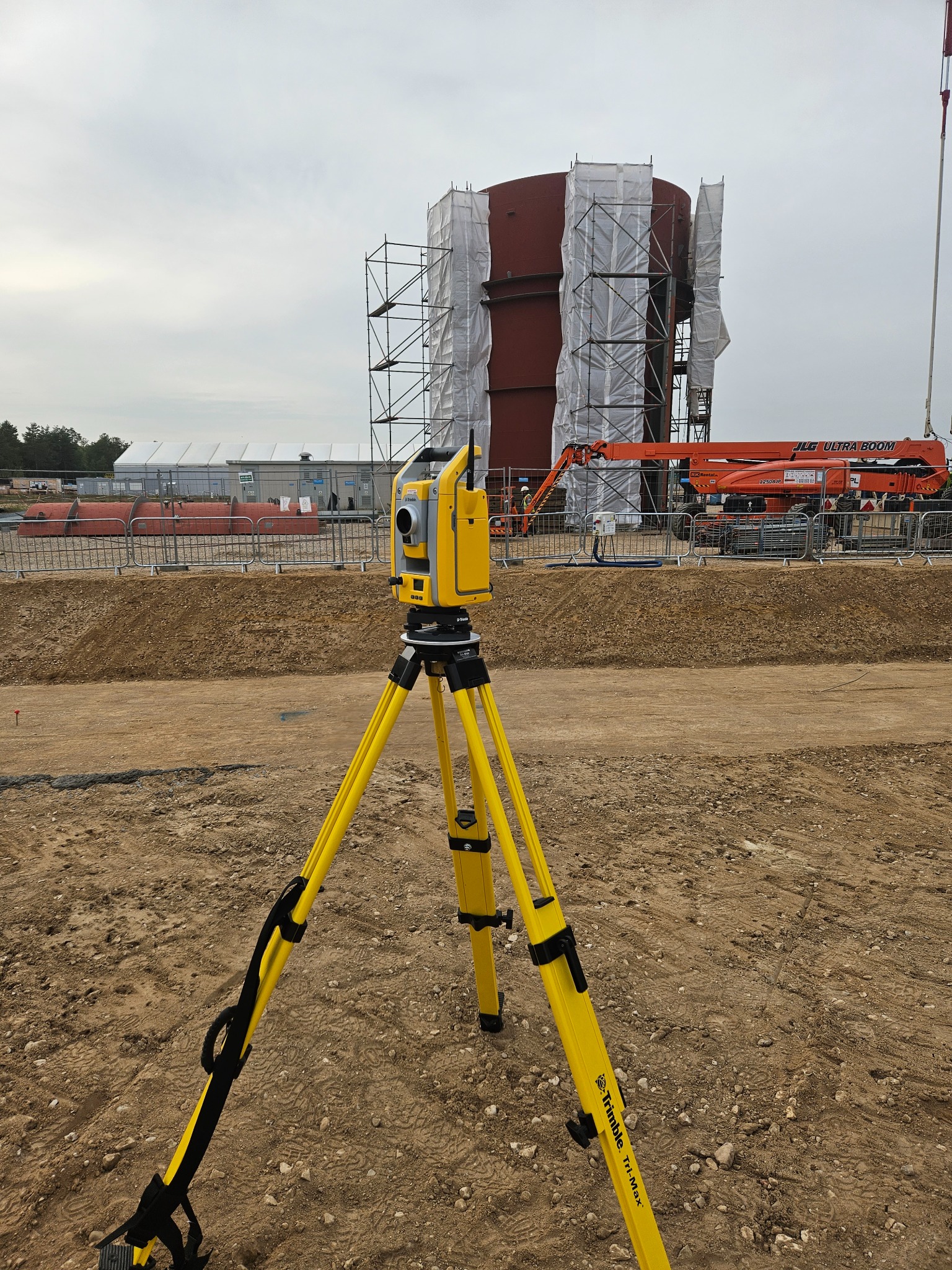 Żółty teodolit marki Trimble Tri-Max na placu budowy z widocznym zbiornikiem w rusztowaniach i podnośnikiem koszowym JLG Ultra Boom w tle.