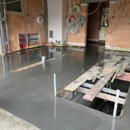 Świeżo wylana, gładka posadzka w trakcie realizacji z widocznymi fragmentami szalunków i instalacji elektrycznej na ścianach z cegły.