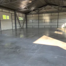 Wnętrze hali przemysłowej z nową, gładką, szarą posadzką betonową, oświetloną naturalnym światłem wpadającym przez okna i tworzącym wyraźny cień.