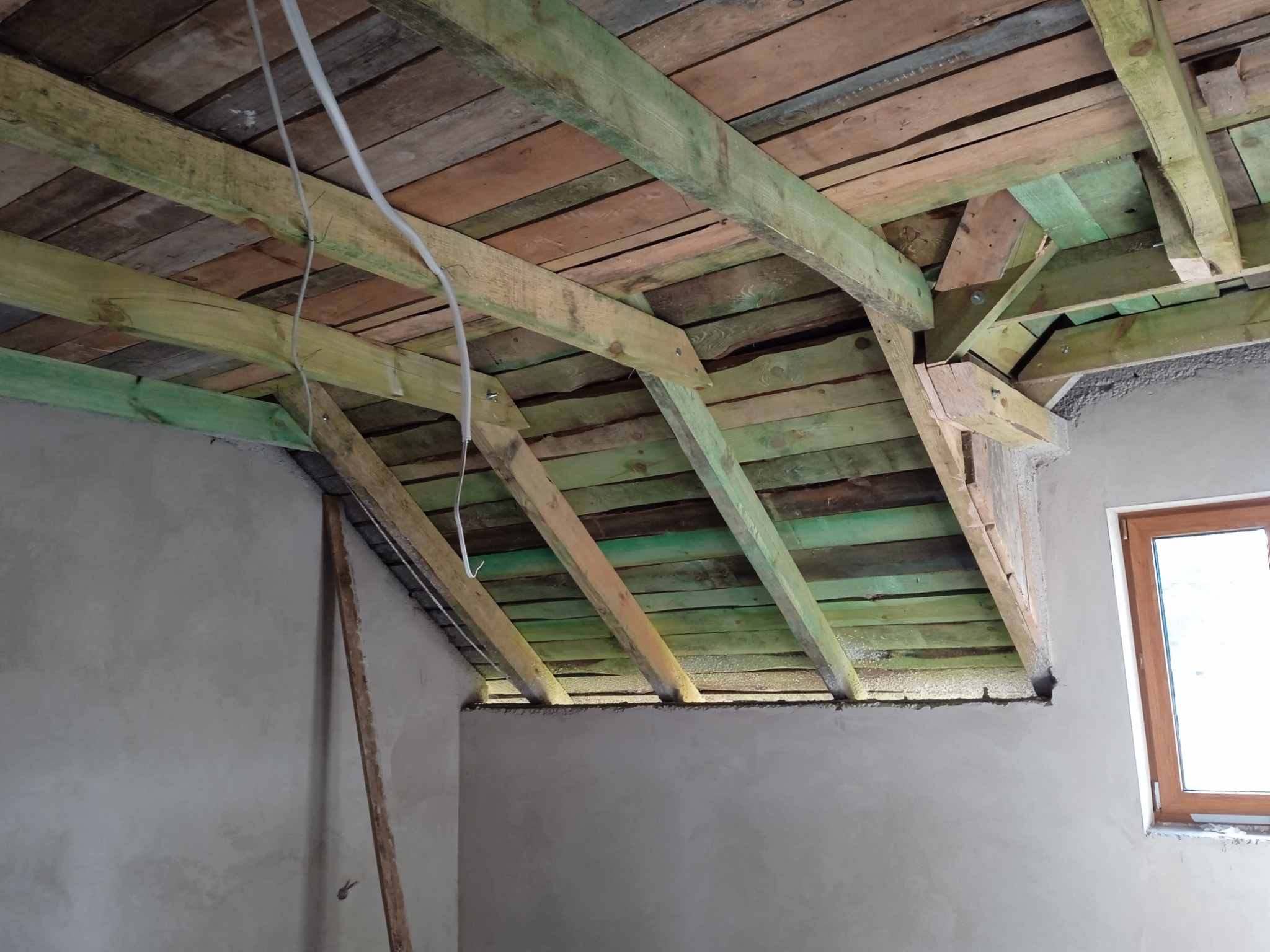 Konstrukcja drewniana dachu z widocznymi belkami, częściowo pomalowanymi na zielono, w surowym wnętrzu z oknem i nieotynkowanymi ścianami.