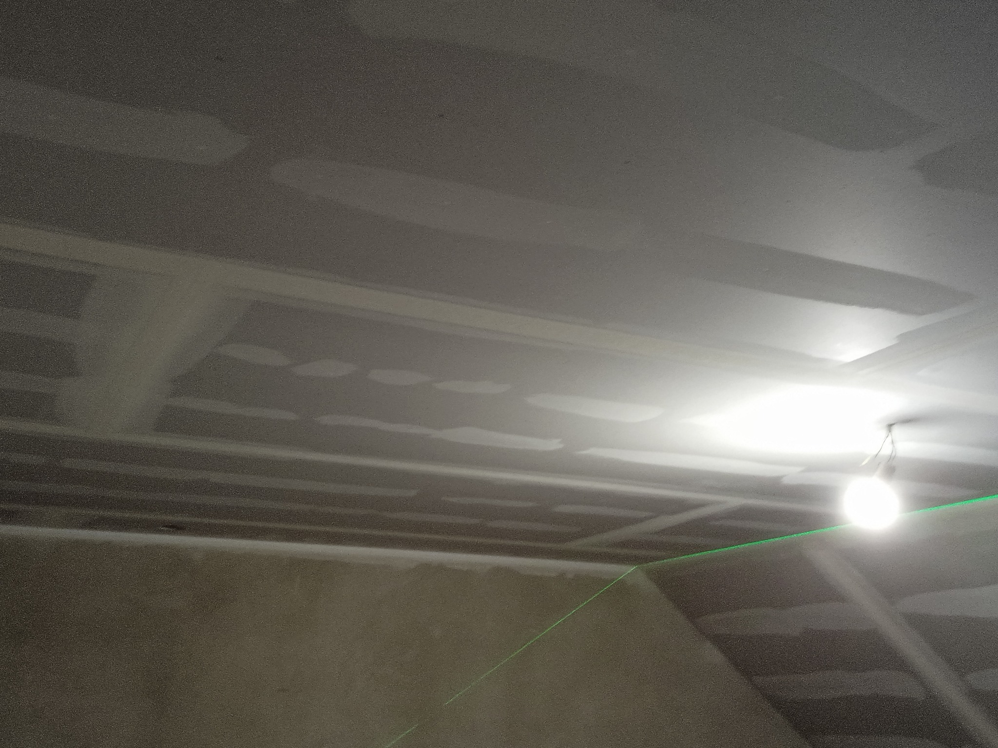Sufit z płyt kartonowo-gipsowych w trakcie szpachlowania, widoczna lampa oświetlająca obszar roboczy i zielona linia lasera wyznaczająca poziom.