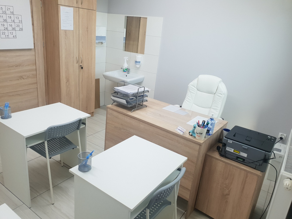 Jasny gabinet z dwoma biurkami, krzesłami z ażurowym oparciem, szafą, umywalką z lustrem i białym fotelem biurowym na kółkach.