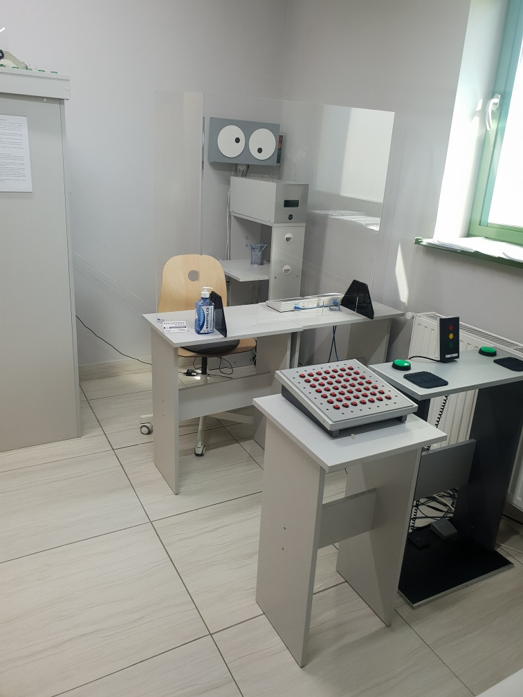 Wnętrze pracowni z aparaturą do badań psychotechnicznych: stanowisko z panelem przycisków, urządzenie z 'oczami', semafor świetlny i pulpit z przyciskami reakcji, oddzielone pleksiglasem...