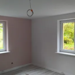 Pomalowane na różowo i biało wnętrze pokoju z dwoma oknami z widokiem na zieleń, podłoga wyłożona panelami imitującymi drewno, widoczne wystające kable ze stropu.