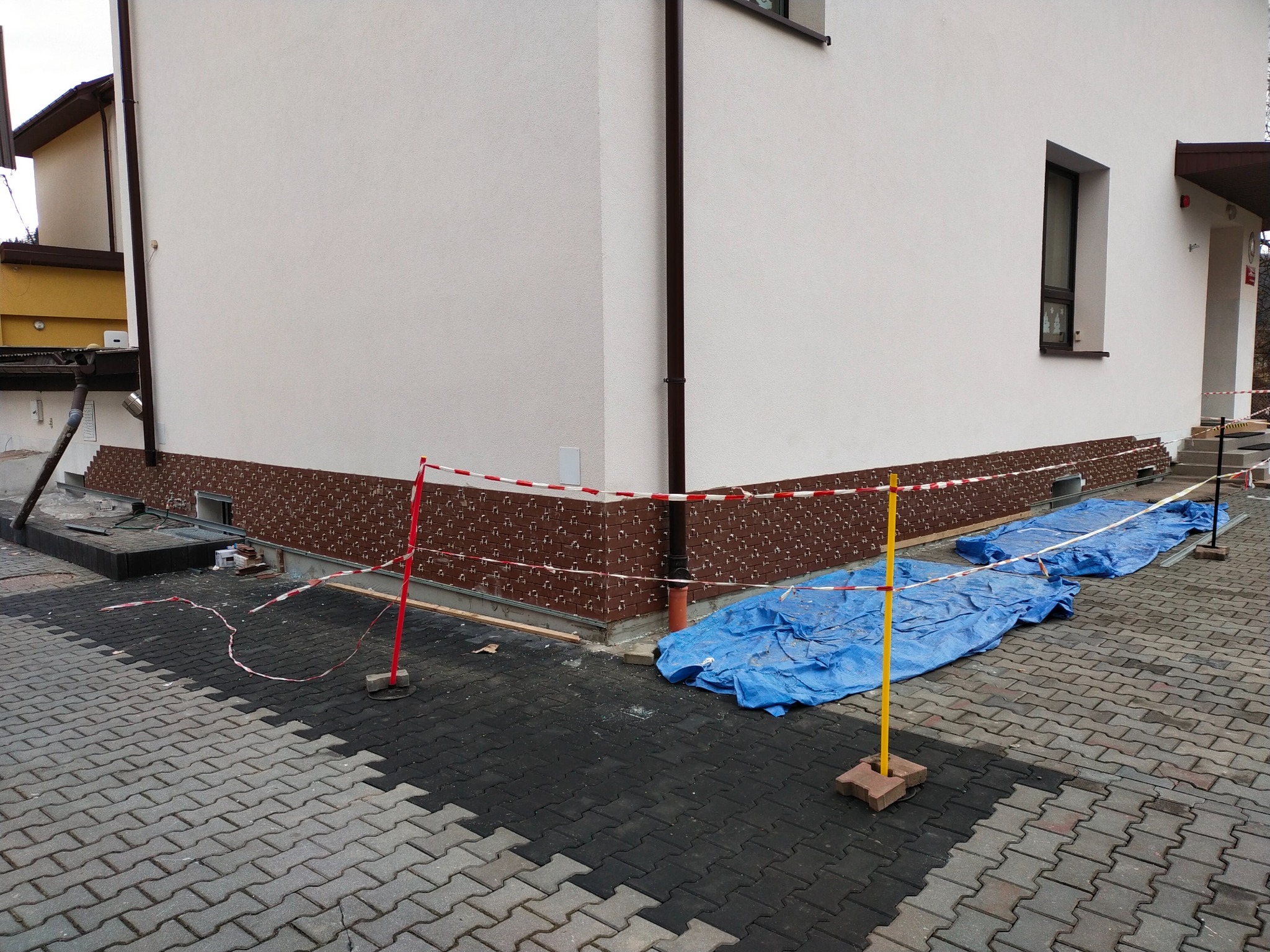 Budynek z częściowo wykończoną elewacją, zabezpieczony folią i taśmą ostrzegawczą, widoczne fragmenty ocieplenia w dolnej części ściany.