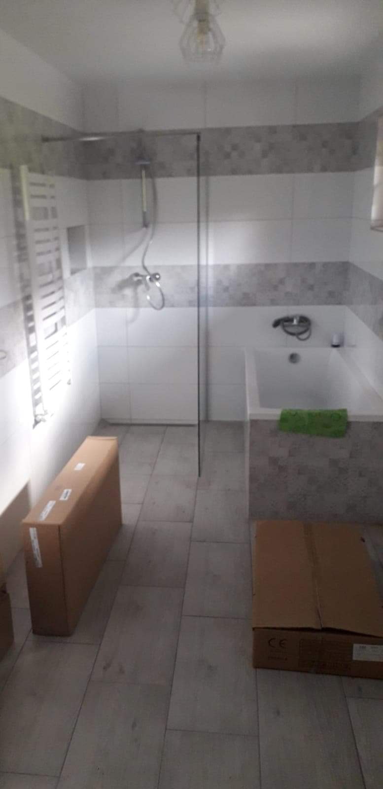 Wnętrze łazienki z wanną, kabiną prysznicową typu walk-in, grzejnikiem drabinkowym i kartonami na podłodze, ukazujące etap prac wykończeniowych.