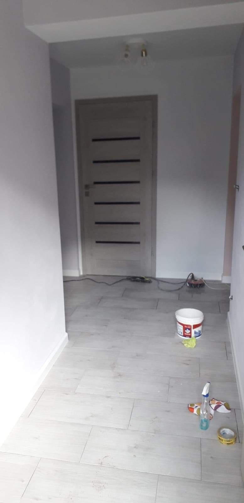 Wnętrze w trakcie remontu w Nowym Sączu: widoczne świeżo pomalowane ściany na biało, jasna podłoga z płytek imitujących drewno, drzwi wewnętrzne z poziomymi wstawkami, puszka z farbą, narzędzia...