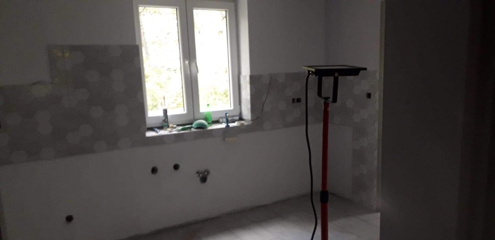 Wnętrze łazienki w trakcie prac wykończeniowych, widoczne fragmenty ściany z szarymi płytkami o heksagonalnym wzorze, przygotowane instalacje wodno-kanalizacyjne oraz narzędzia na parapecie okna.