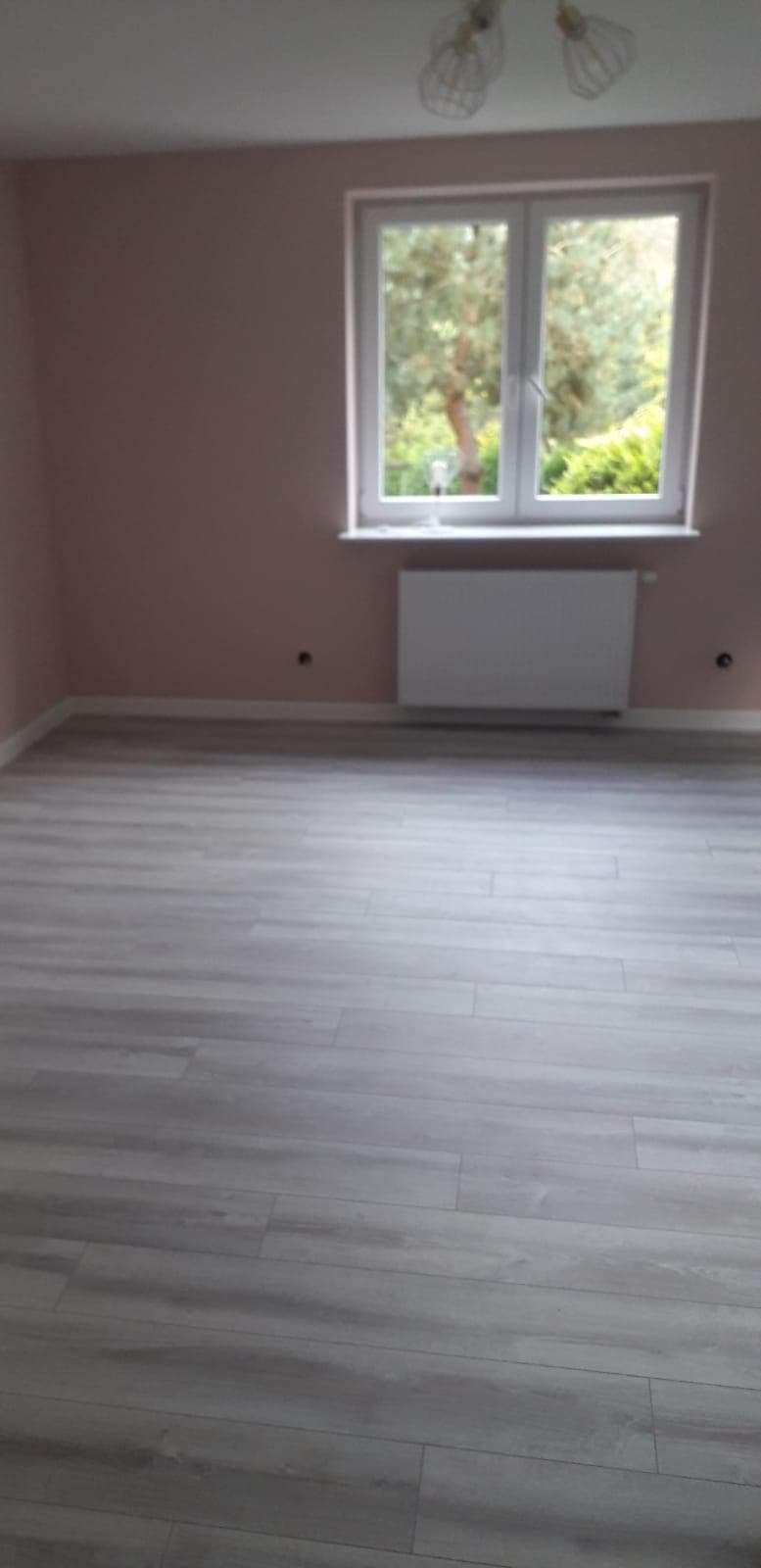 Jasnoszara podłoga imitująca drewno w nowo wykończonym pokoju z różowymi ścianami, białą listwą przypodłogową i grzejnikiem pod oknem.