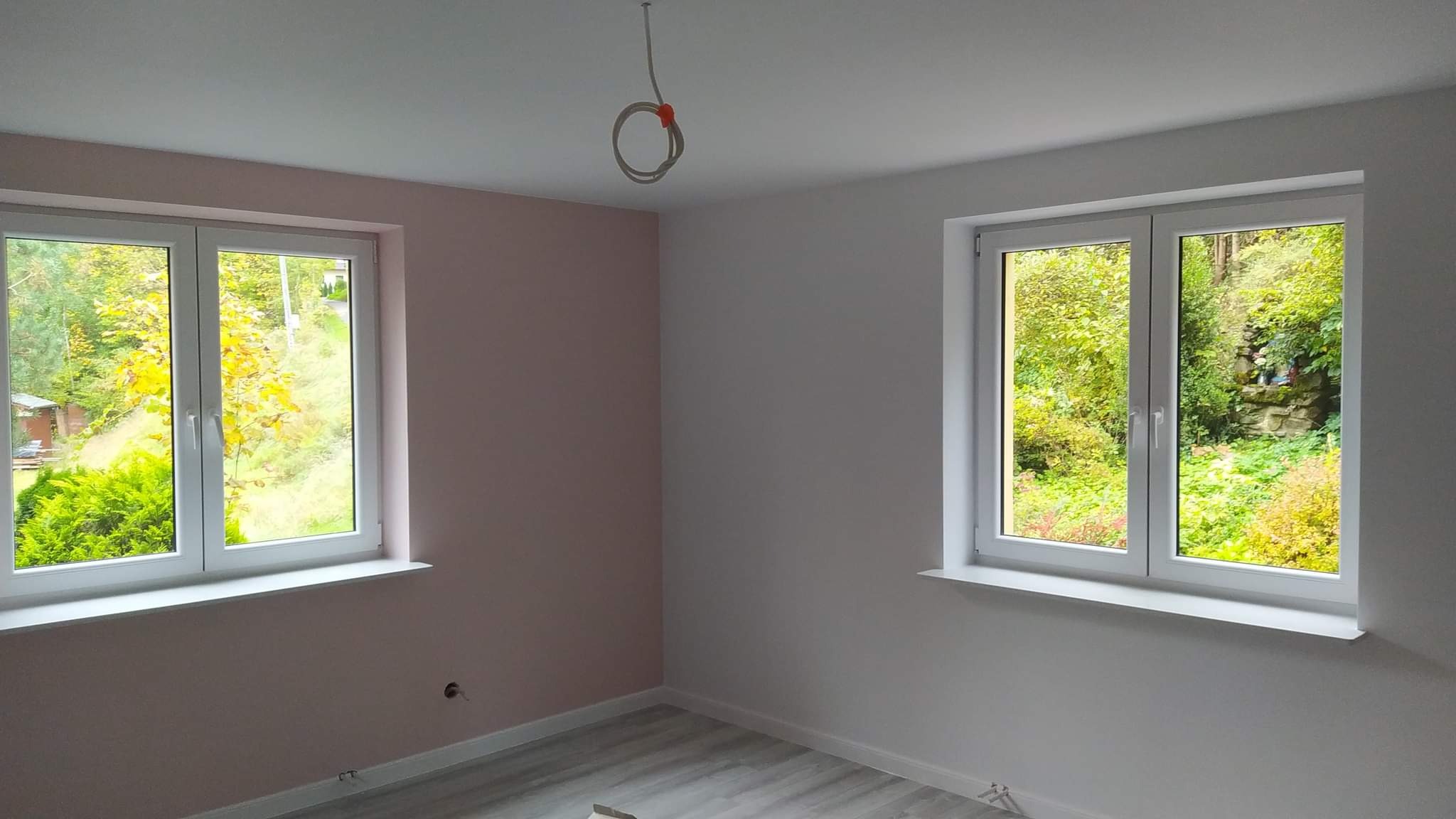 Pomalowane na różowo i biało wnętrze pokoju z dwoma oknami z widokiem na zieleń, podłoga wyłożona panelami imitującymi drewno, widoczne wystające kable ze stropu.