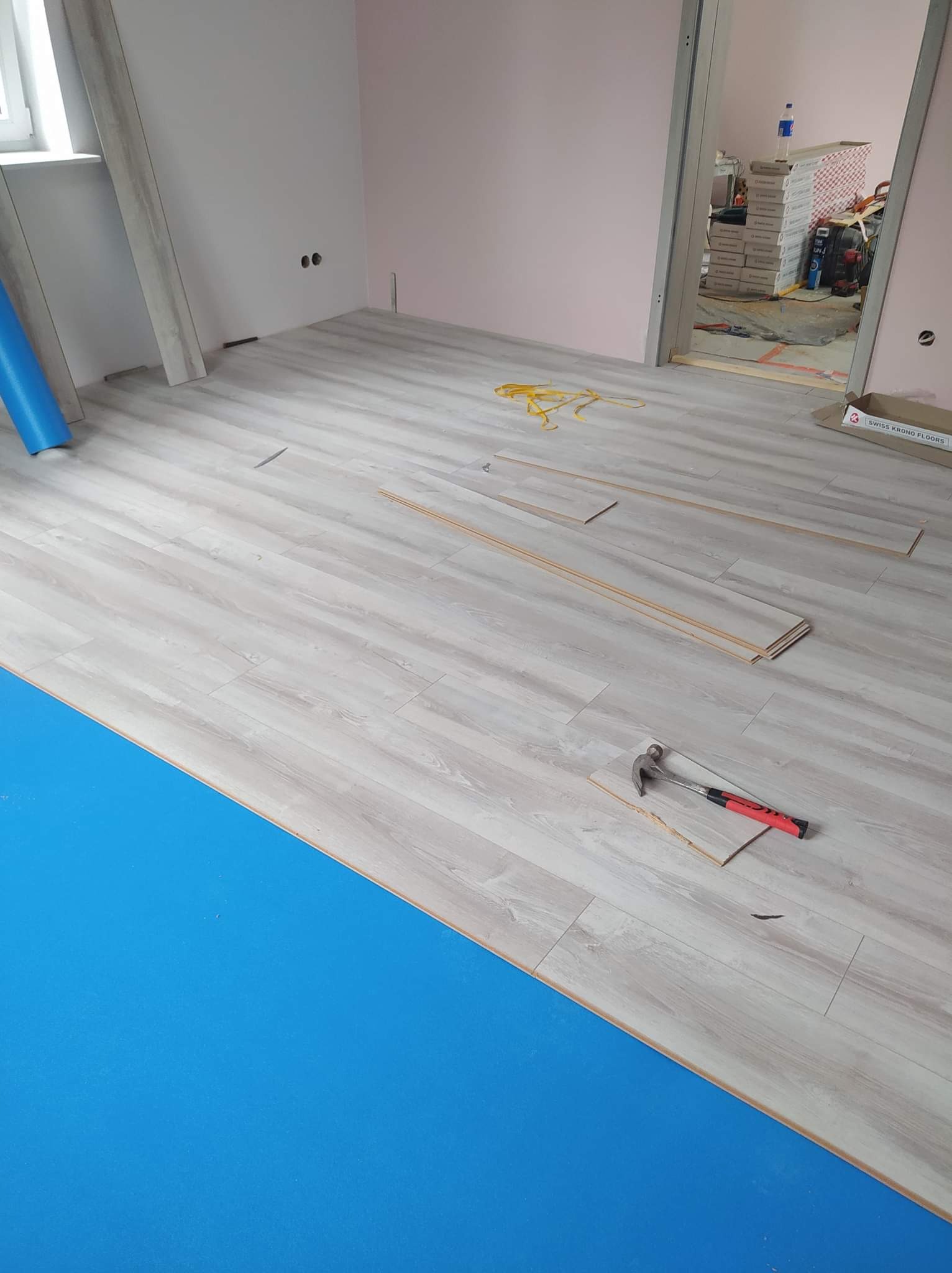 Układanie paneli podłogowych: widoczne panele Swiss Krono Floors, niebieska mata pod panele, młotek z czerwonym uchwytem, częściowo ułożona podłoga w jasnym odcieniu drewna, żółta miarka...
