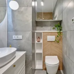 Nowoczesna łazienka z szarą marmurową glazurą, owalną białą umywalką nablatową z baterią w kolorze złotym, okrągłym lustrem z podświetleniem LED, drewnianą ścianą w jodełkę i wiszącą toaletą.