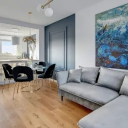 Nowoczesne wnętrze mieszkania w odcieniach bieli, szarości i niebieskiego z drewnianą podłogą, obejmujące otwartą kuchnię z białymi szafkami, jadalnię ze szklanym stołem i czarnymi krzesłami...