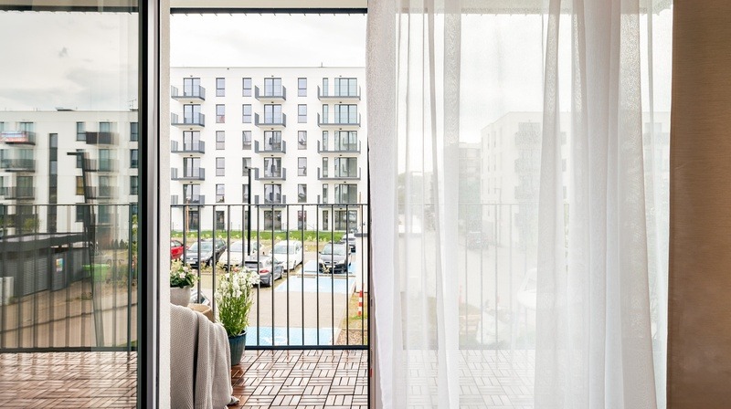 Widok z balkonu na nowe osiedle: drewniana podłoga balkonu, czarne barierki, kwiaty w donicach, częściowo zasłonięte firanką, w tle parking i budynki mieszkalne.