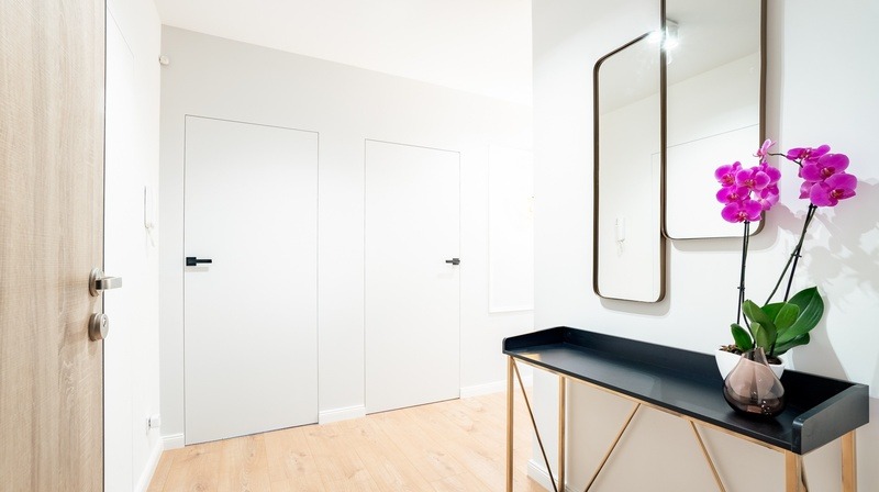 Jasne wnętrze przedpokoju z dwiema białymi, bezprzylgowymi drzwiami z czarnymi klamkami, drewnianymi drzwiami z lewej strony, lustrem w metalowej ramie i konsolą z fioletowymi storczykami.
