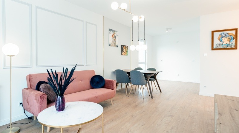 Jasne wnętrze mieszkania z parkietem, różową sofą, okrągłym stolikiem z marmurowym blatem, lampą podłogową z kulistym kloszem i designerską lampą sufitową z kulistymi kloszami, dekoracyjnymi...