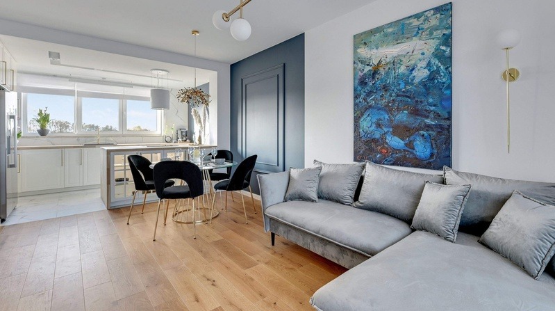 Nowoczesne wnętrze mieszkania w odcieniach bieli, szarości i niebieskiego z drewnianą podłogą, obejmujące otwartą kuchnię z białymi szafkami, jadalnię ze szklanym stołem i czarnymi krzesłami...