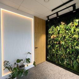 MAJSTER PRO TEAM - Nowoczesne wnętrze biurowe z pionową ścianą zieleni w czarnej ramie, oświetloną ścianą z białymi lamelami z ledowym podświetleniem, drewnianymi panelami i roślinami doniczkowymi, tworzącymi...