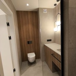 MAJSTER PRO TEAM - Nowoczesna łazienka z zabudową meblową z drewna, wiszącą toaletą, umywalką z szafką i dekoracyjnym oświetleniem, widok zza lustra.