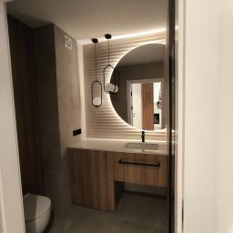 MAJSTER PRO TEAM - Nowoczesna łazienka z okrągłym lustrem podświetlanym LED, drewnianą szafką pod umywalką i wiszącymi lampami o geometrycznym kształcie.