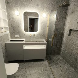 MAJSTER PRO TEAM - Nowoczesna łazienka w trakcie montażu: szare płytki lastryko na ścianach i podłodze, podświetlane lustro z zaokrąglonymi rogami, biała szafka pod umywalkę z otwartą szufladą, kabina prysznicowa ze...