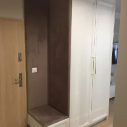 Wnęka z tapicerowanym siedziskiem i panelem obok drzwi wejściowych, zintegrowana z białą szafą w przedpokoju, drewniana podłoga.