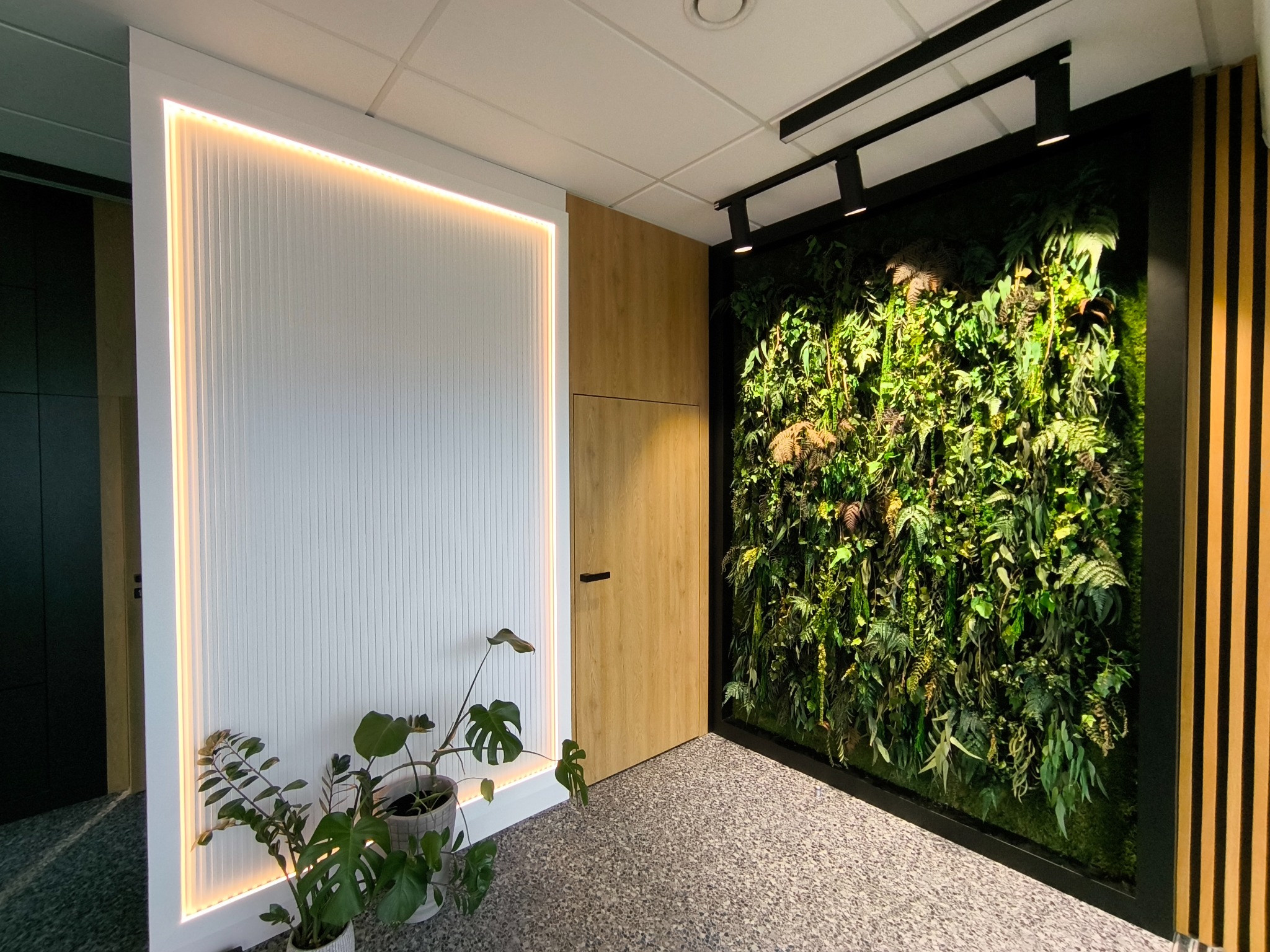 Nowoczesne wnętrze biurowe z pionową ścianą zieleni w czarnej ramie, oświetloną ścianą z białymi lamelami z ledowym podświetleniem, drewnianymi panelami i roślinami doniczkowymi, tworzącymi...