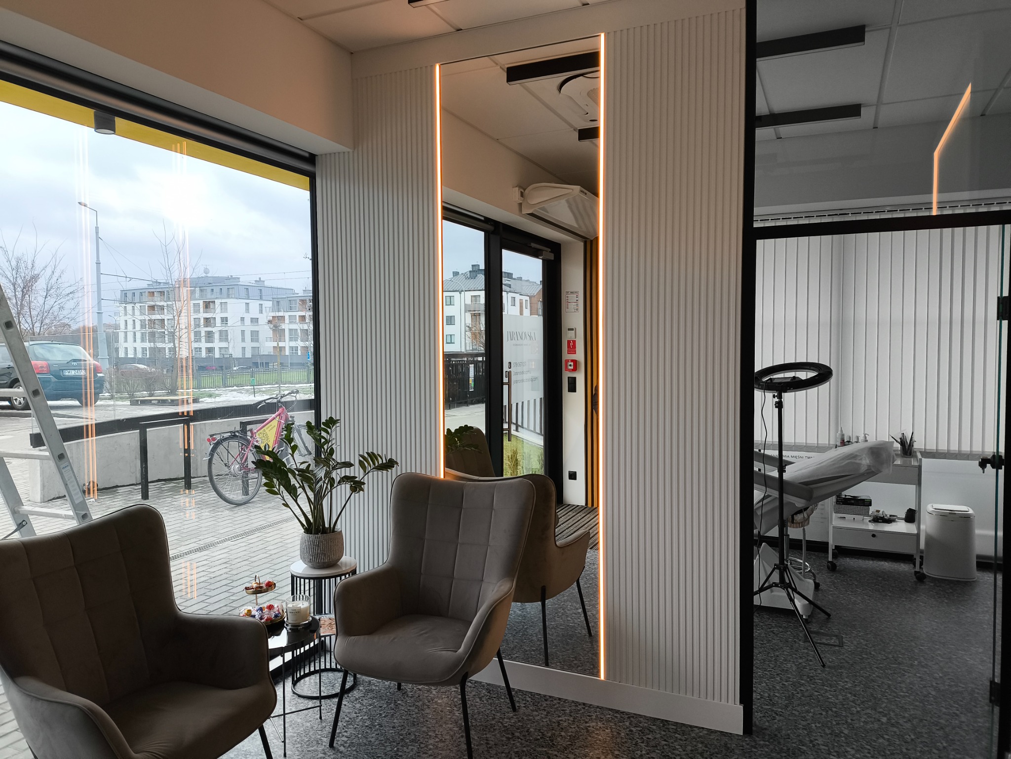 Wnętrze gabinetu kosmetycznego z dwoma fotelami, stolikiem kawowym i lustrem z podświetleniem LED, widok na ulicę przez okno i stanowisko zabiegowe w tle.
