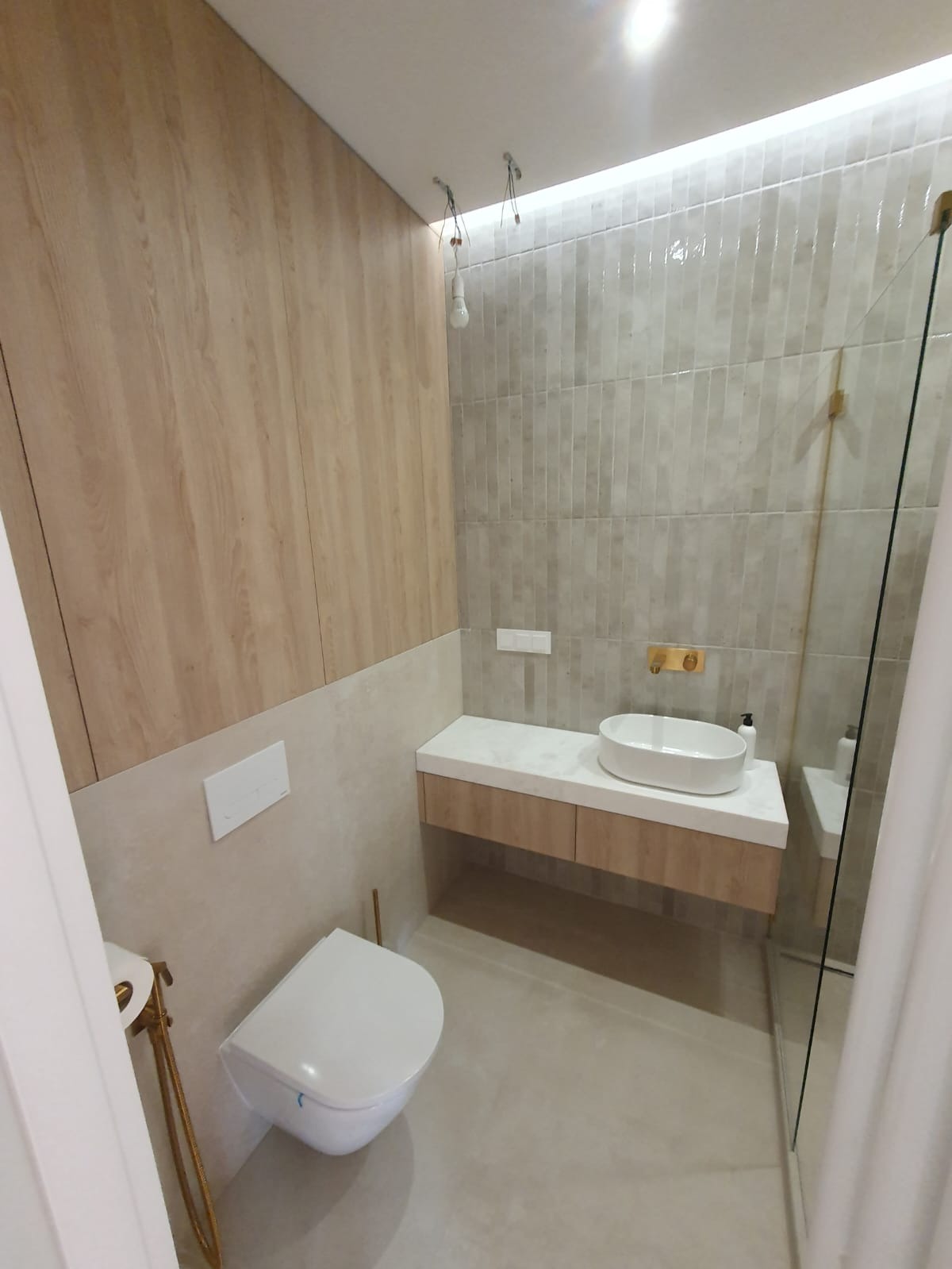 Nowoczesna łazienka z drewnianą okładziną ścienną, płytkami w odcieniach szarości, podwieszaną toaletą i umywalką nablatową na szafce z blatem imitującym marmur, widoczne złote akcenty.
