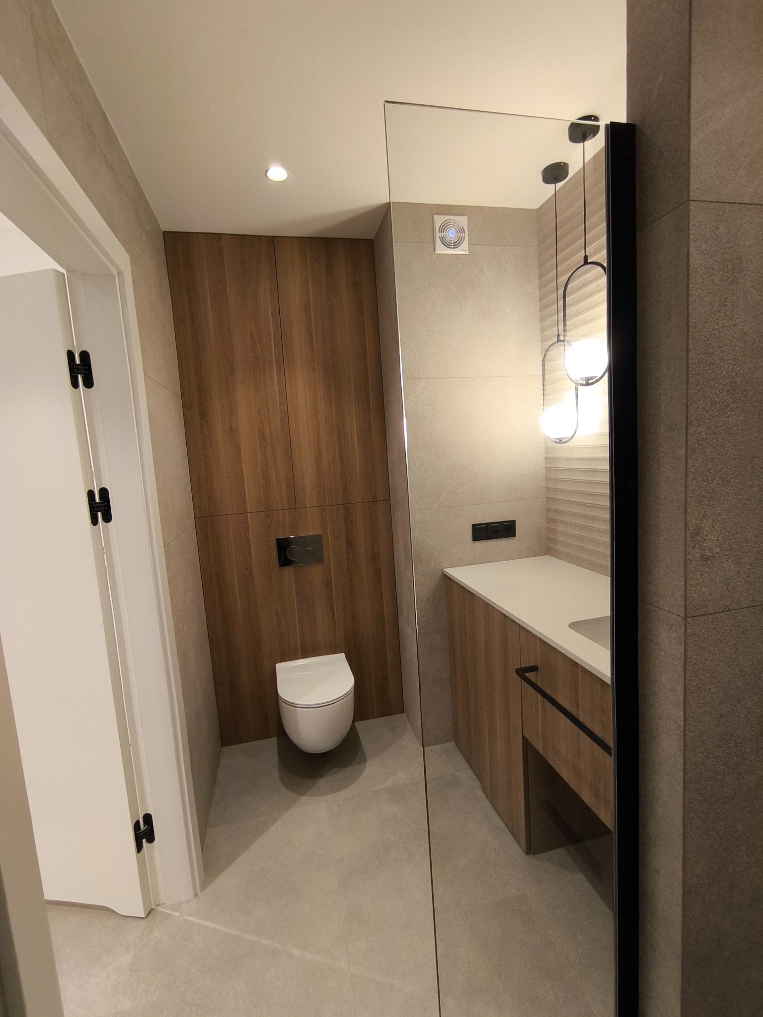 Nowoczesna łazienka z zabudową meblową z drewna, wiszącą toaletą, umywalką z szafką i dekoracyjnym oświetleniem, widok zza lustra.