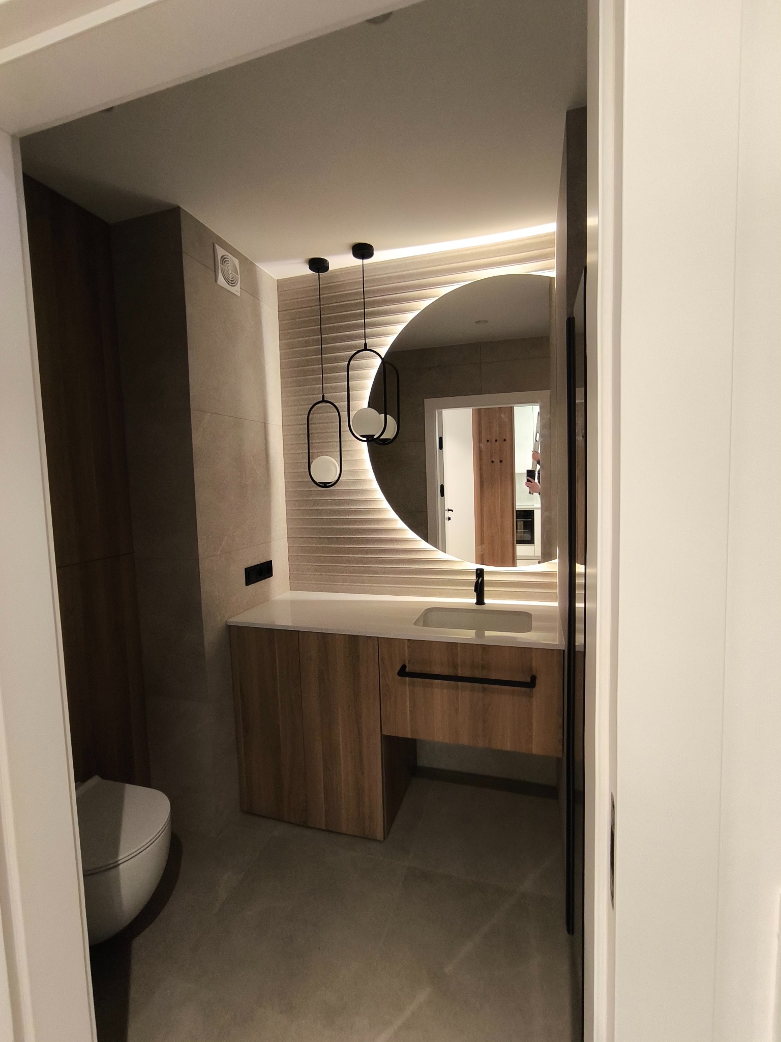 Nowoczesna łazienka z okrągłym lustrem podświetlanym LED, drewnianą szafką pod umywalką i wiszącymi lampami o geometrycznym kształcie.