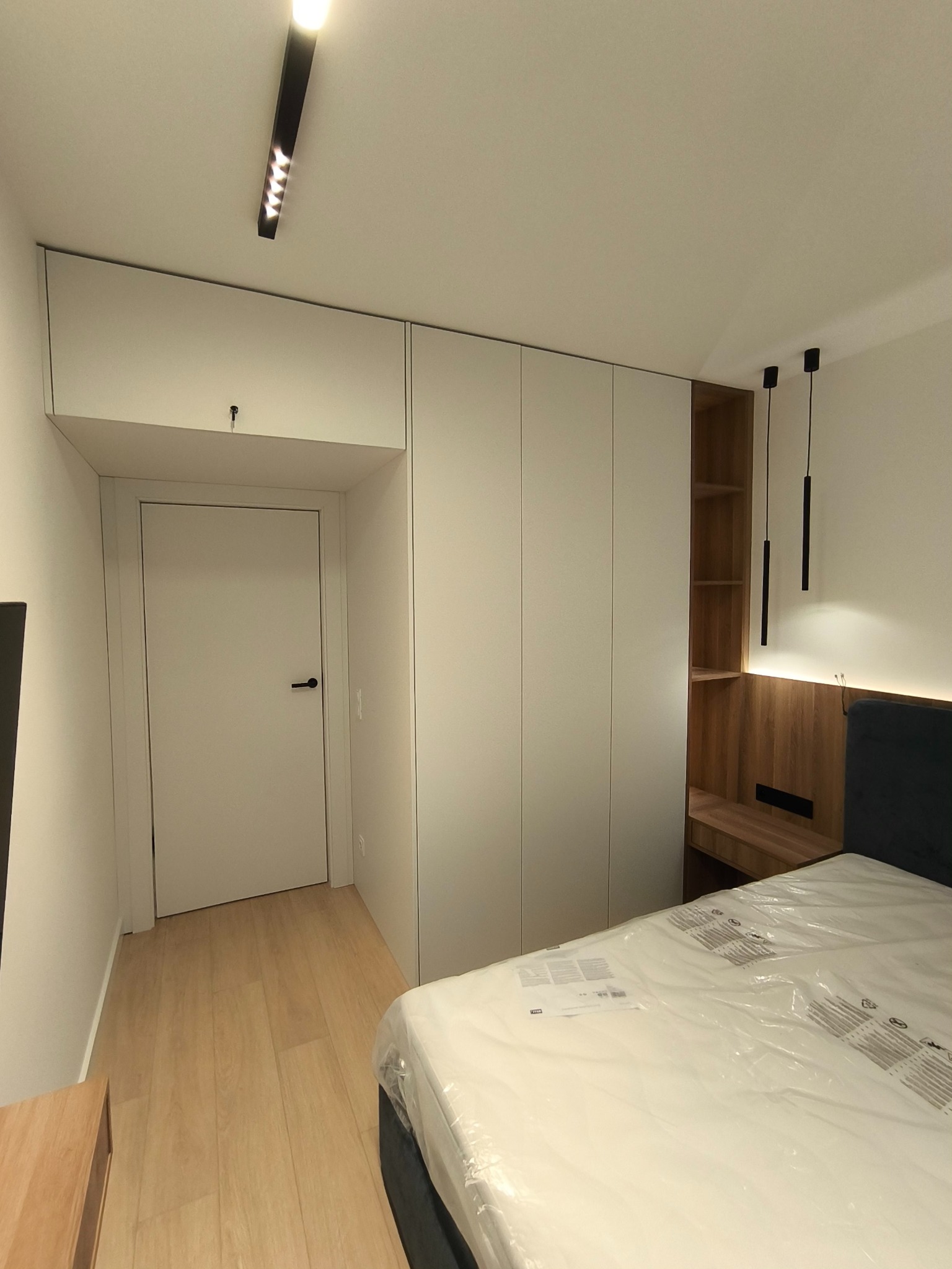 Nowoczesna sypialnia z zabudową meblową: białe szafy wnękowe z drzwiami bez uchwytów, drewniana wnęka z półkami i oświetleniem LED nad wezgłowiem łóżka, minimalistyczne czarne lampy wiszące...