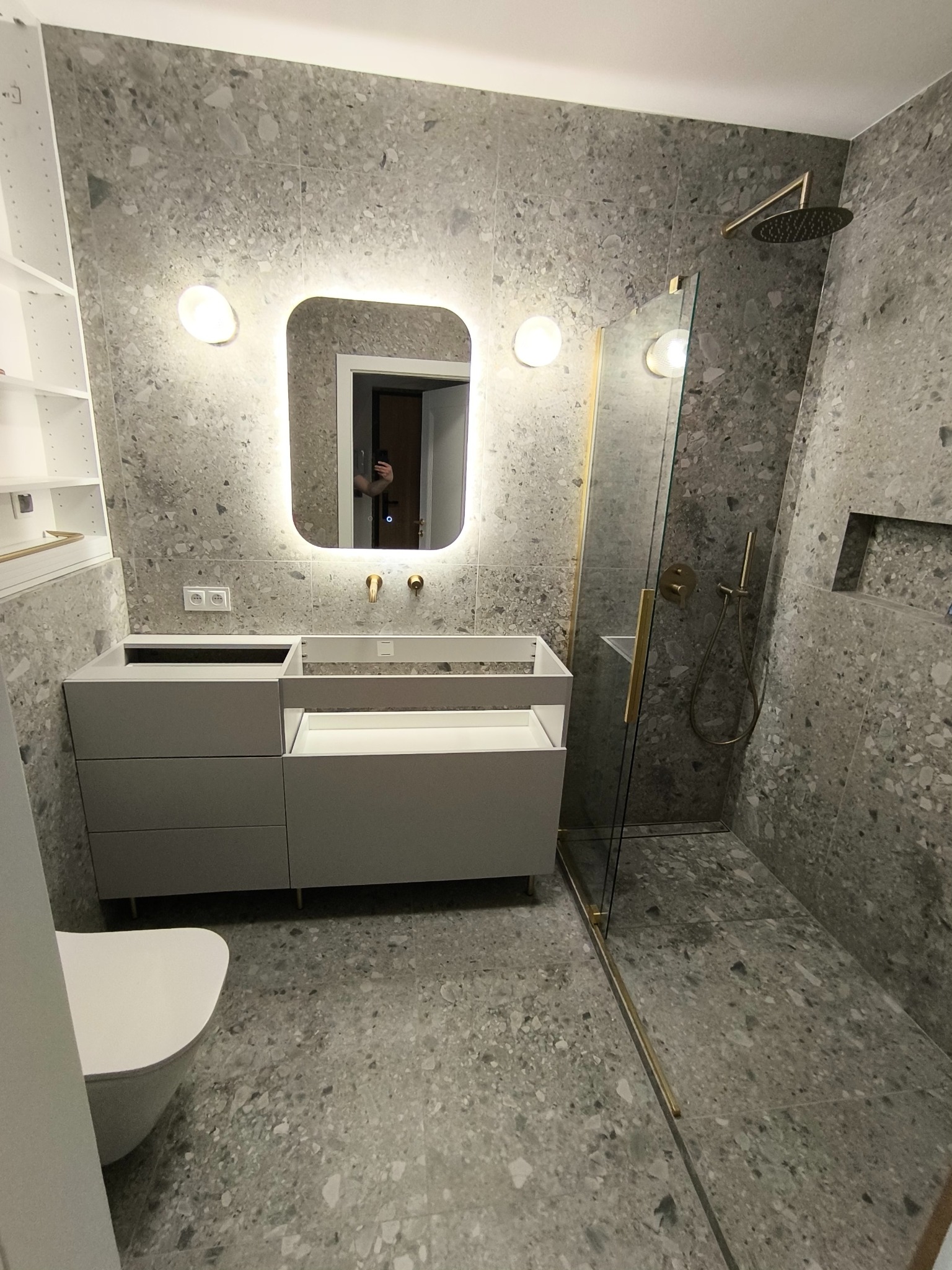 Nowoczesna łazienka w trakcie montażu: szare płytki lastryko na ścianach i podłodze, podświetlane lustro z zaokrąglonymi rogami, biała szafka pod umywalkę z otwartą szufladą, kabina prysznicowa ze...