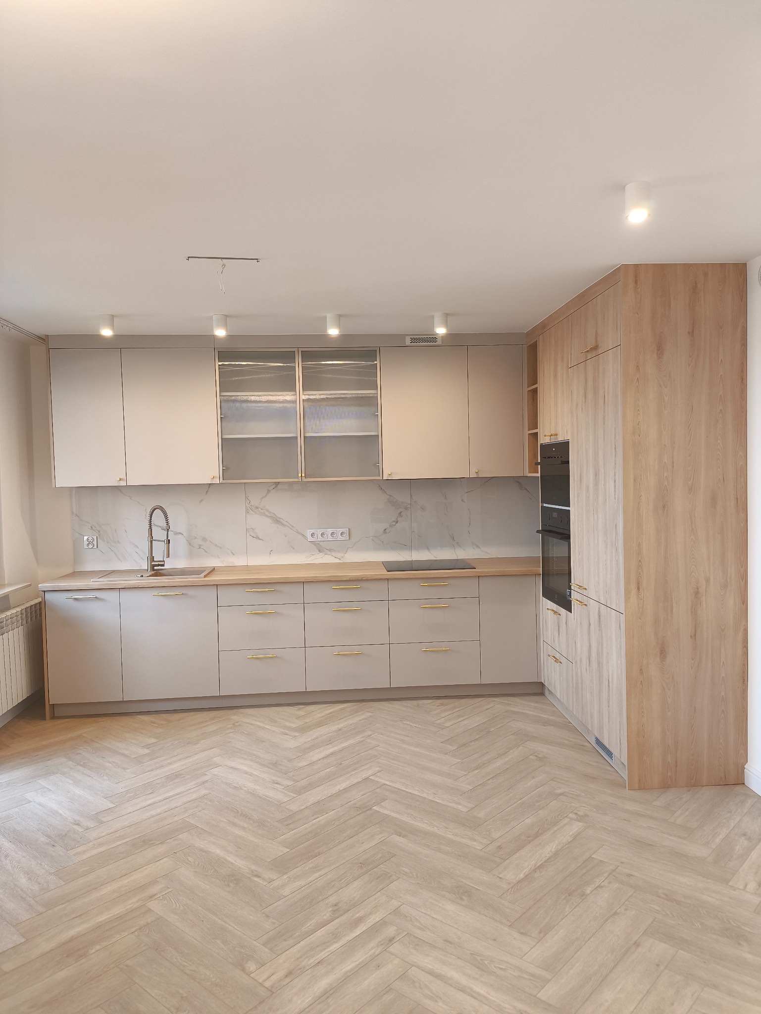 Jasna kuchnia w stylu skandynawskim z drewnianą podłogą ułożoną w jodełkę, beżowe szafki z złotymi uchwytami, marmurowy backsplash i nowoczesne oświetlenie sufitowe.